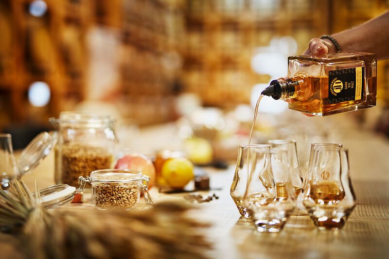 A hand pours whisky from a bottle into several glasses on a table with cereals and fruit.