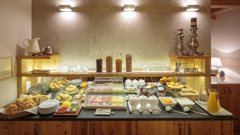 A breakfast buffet with bread rolls, cold cuts, muesli, fruit and drinks on a wooden counter.