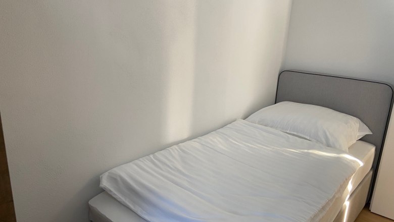A simple, neatly made single bed in a bright room with white walls.