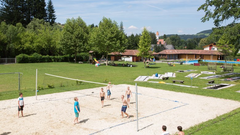 Children play volleyball on a sand court at the Aspang outdoor pool, surrounded by green meadows and trees.
