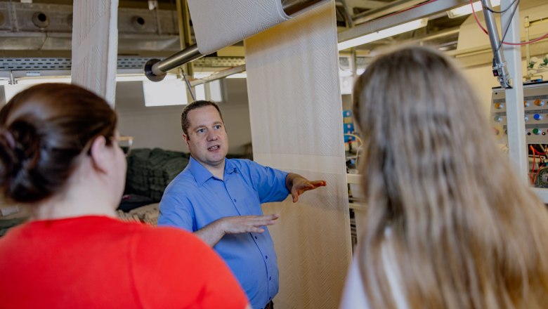 A man in a blue shirt explains something to two women on a factory floor.
