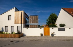 Modern building with a white façade and wooden elements, surrounded by a wall with a wooden gate.