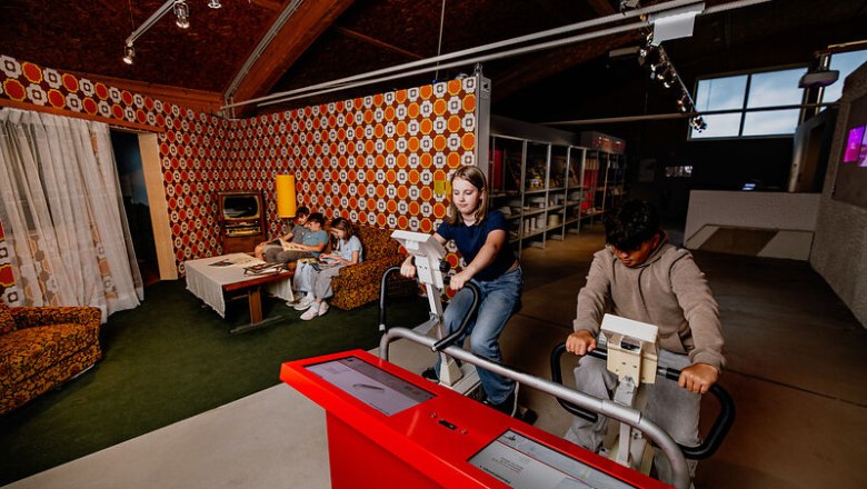 Young people on bicycle ergometers in a room with retro living room decor.