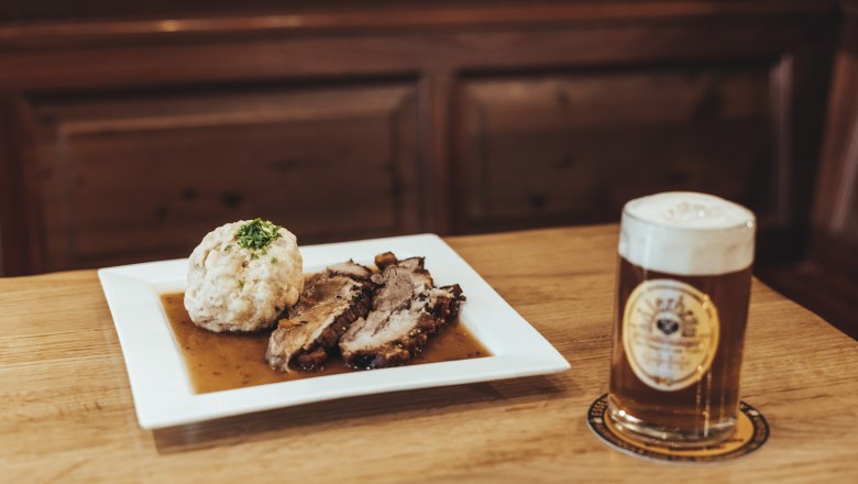 A plate of roast pork and dumplings next to a glass of beer on a wooden table.