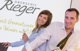 Two people are smiling and holding bottles of wine in front of a sign that reads 'Oberschil Rieger'.