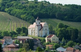 A church in the middle of vineyards and houses, surrounded by green hills.