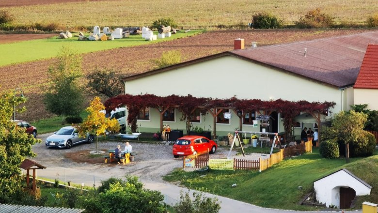 A rural building covered in vines, surrounded by fields and cars. People are sitting at a table outside.