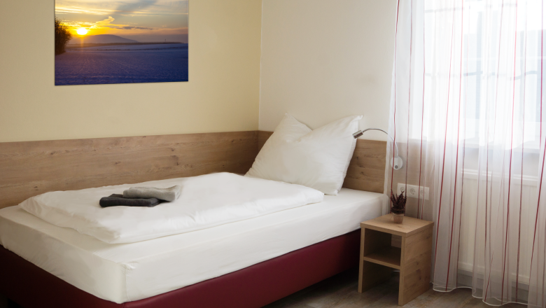 A simple single room with a bed, a bedside table and a picture on the wall. A window with curtains lets in light.