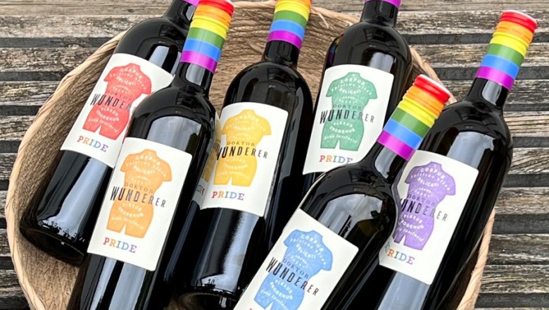 Six wine bottles with rainbow labels in a basket on a wooden table.
