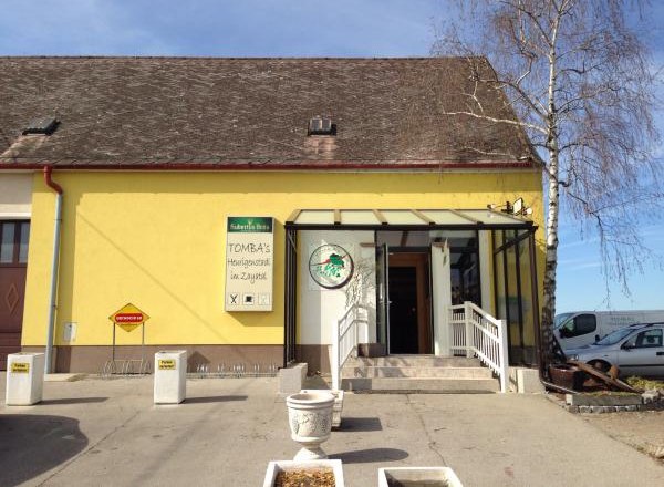 Yellow building with sign 'Tomba's Heurigenschenke'. Entrance with stairs and glass roof.