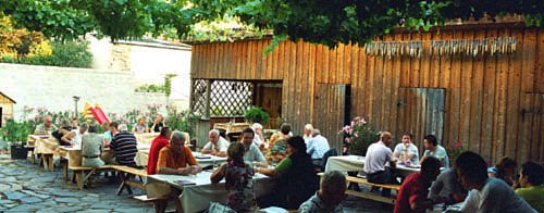 People sit at wooden tables in a cozy courtyard with vines.