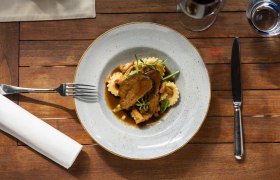 A plate of braised beef cheeks, ravioli and vegetables on a wooden table, with cutlery and a glass of red wine next to it.
