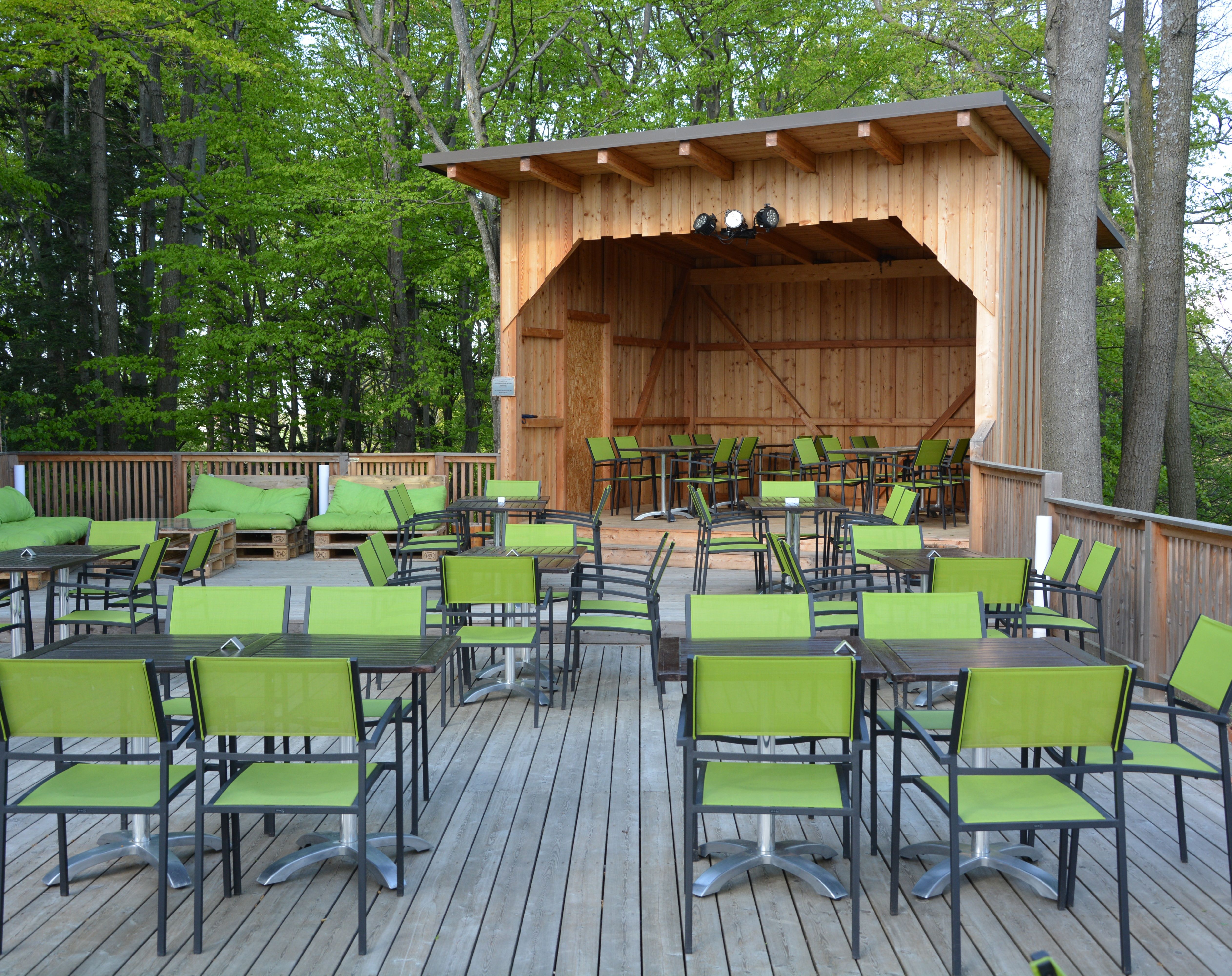A wooden terrace with black tables and green chairs and a covered wooden stage.