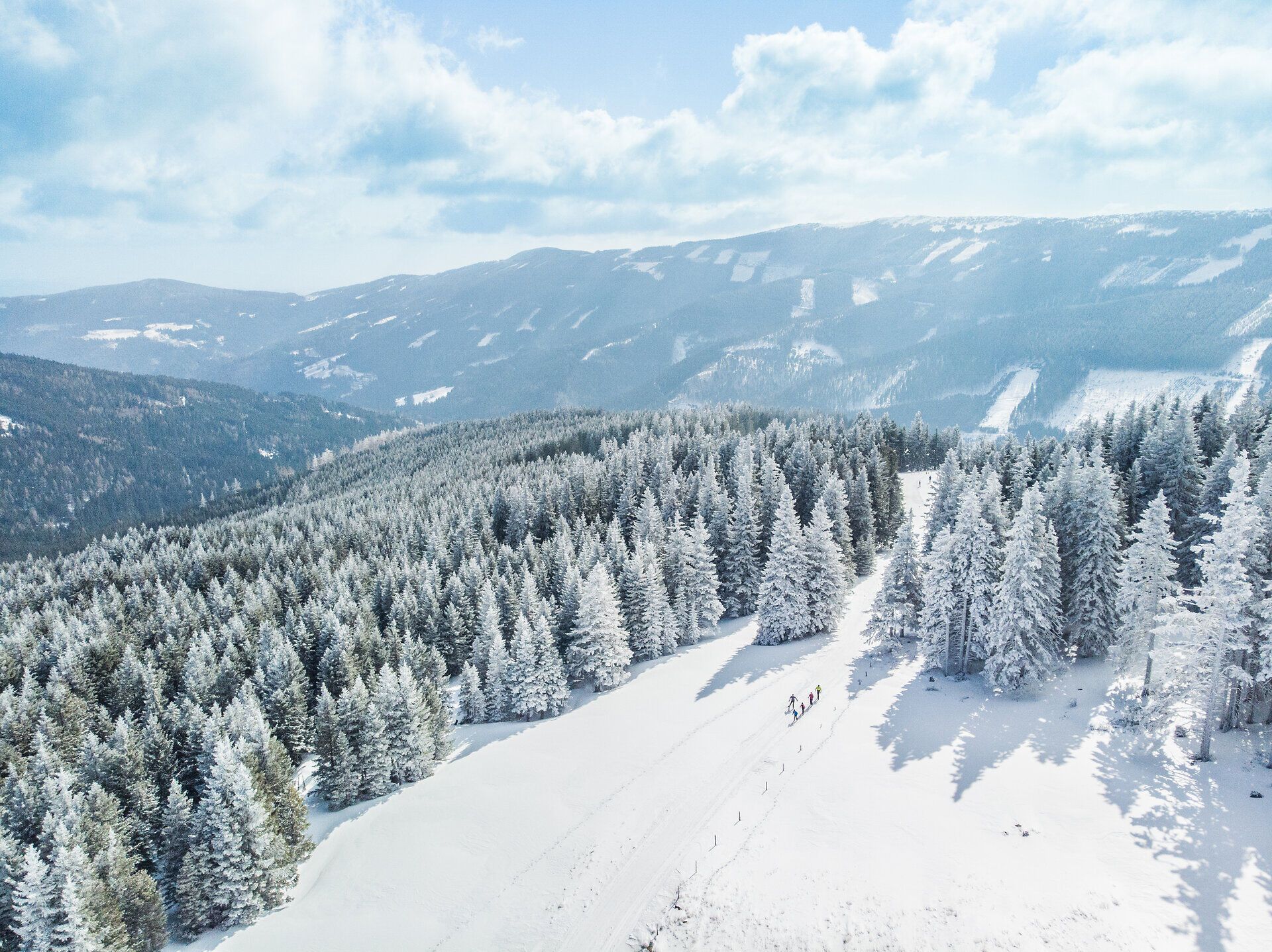 Luftaufnahme der verschneiten Wechsel-Panoramaloipe zwischen schneebedeckten Nadelbäumen mit Blick auf die umliegende Berglandschaft.