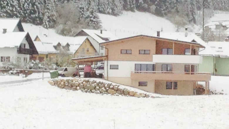 Modern house in the snow with surrounding buildings and forest in the background.