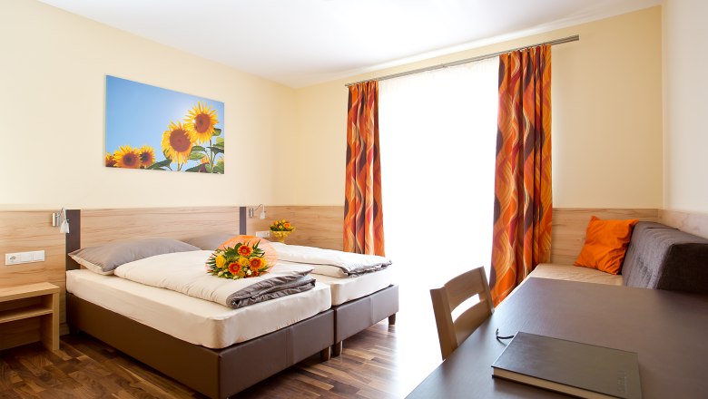 A bright guest room with two beds, a table, a sofa and a picture of sunflowers on the wall.