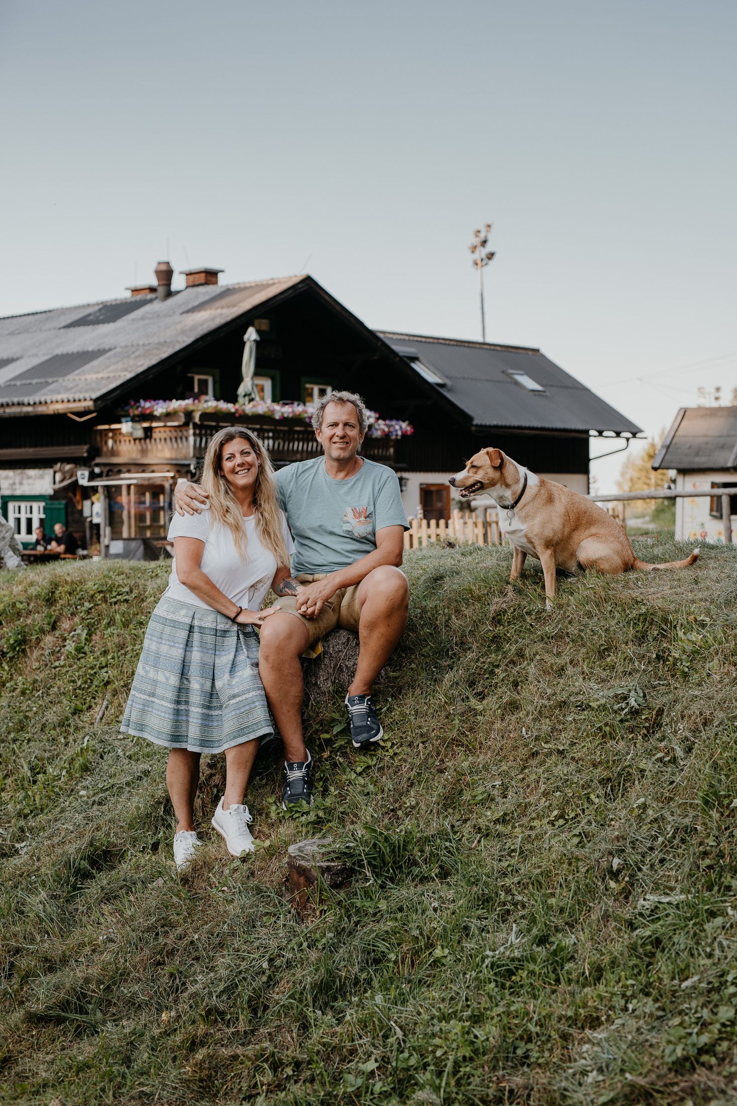A couple sits on a hill in front of a hut with a dog next to them.