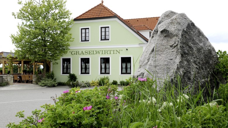 Green building with the inscription 'Graselwirtin', surrounded by plants and a large rock in the foreground.