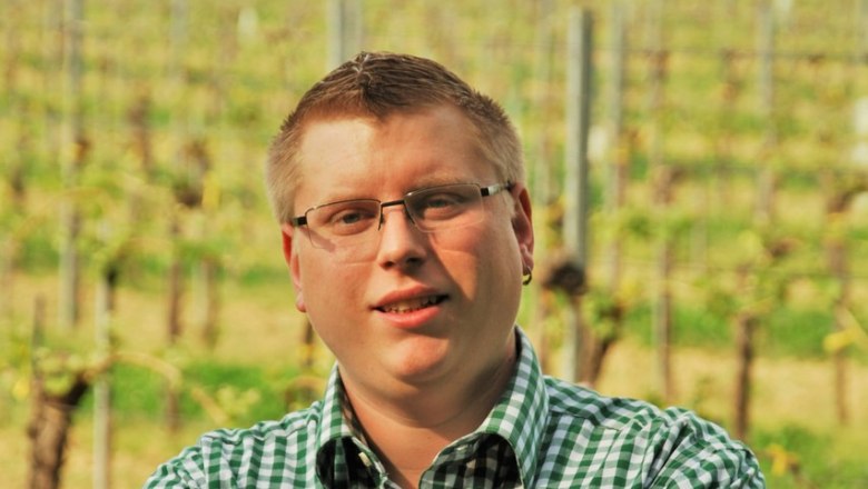 A man with glasses and a checked shirt stands with his arms folded in front of a vineyard.