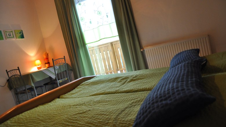 Cozy bedroom with green bed, table and chairs, lamp and window with curtains.