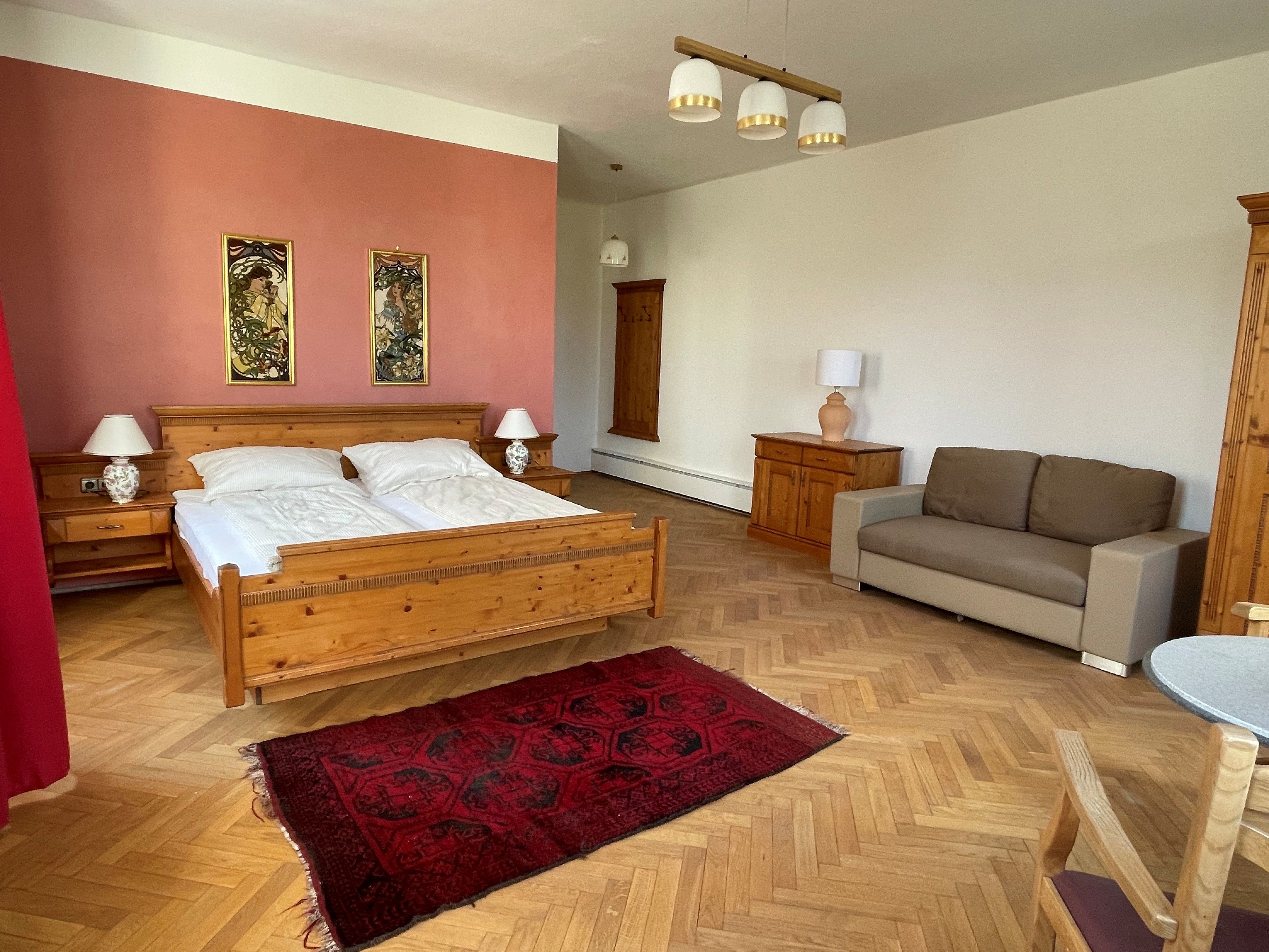 A spacious bedroom with a wooden bed, nightstands, a couch, and a red rug on a wooden floor.