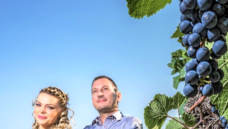 A man and a woman stand in front of a blue sky next to a vine with grapes.