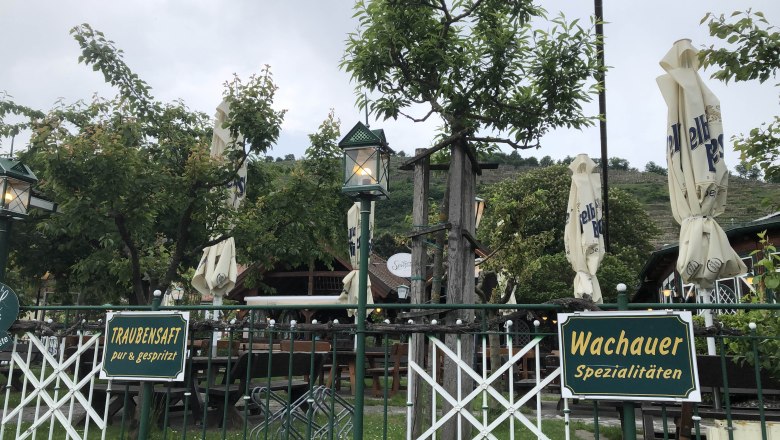 Guest garden with signs for grape juice and Wachau specialties, surrounded by trees and closed parasols.
