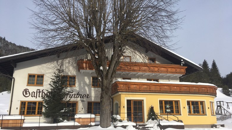 Gasthof Furtner, © Gasthof Furtner An inn in the snow with wooden decorations and a large tree in front of it.