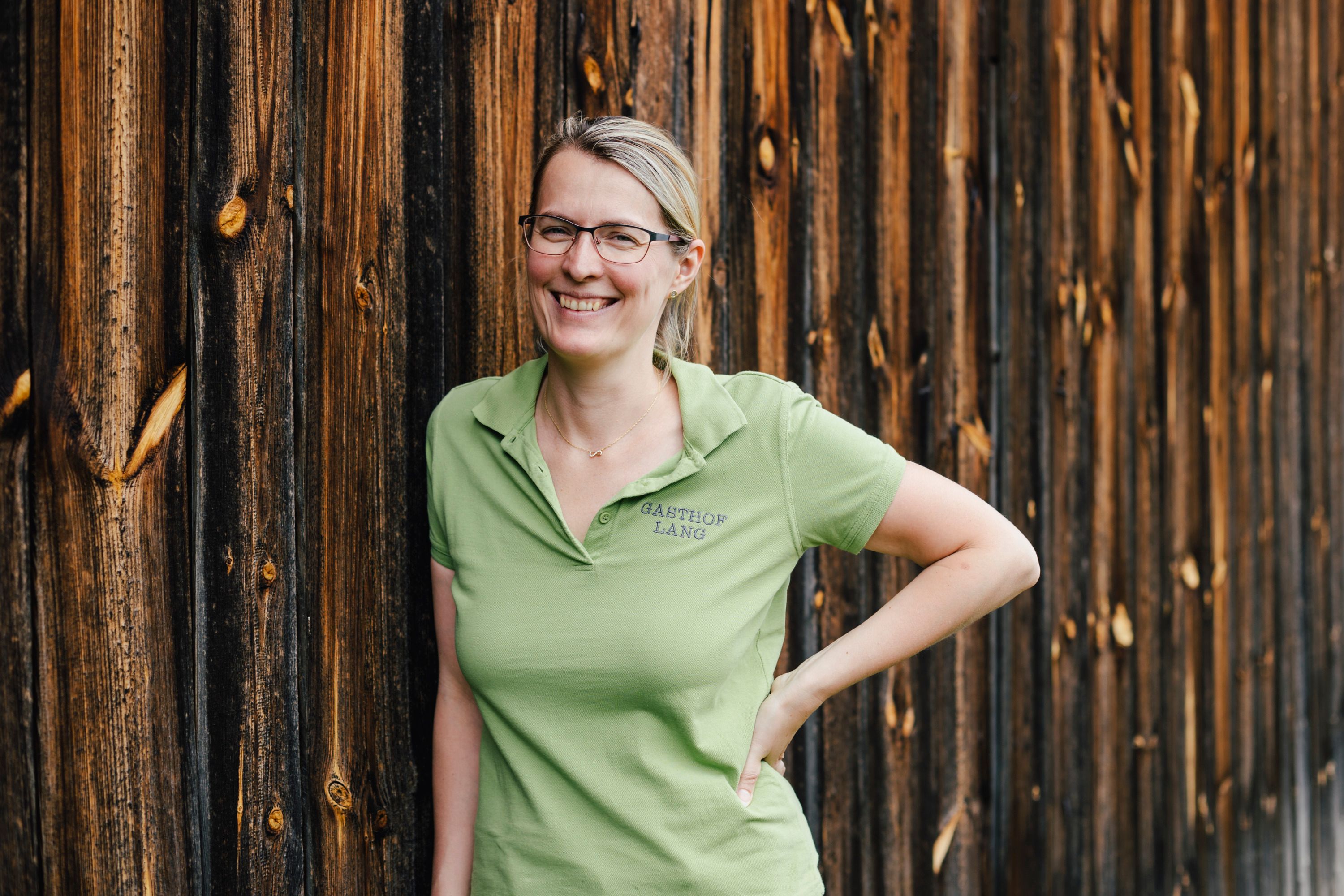 A woman in a green polo shirt leans against a wooden wall, smiling.