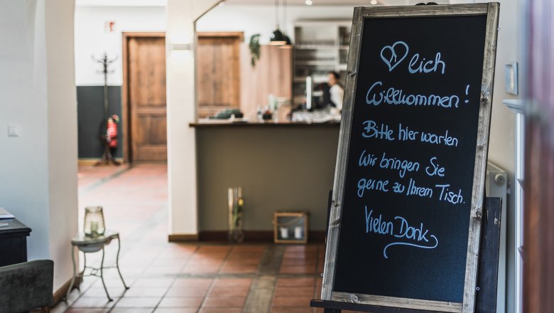 Entrance area of a restaurant with a sign welcoming guests and asking them to wait until they are brought to their table.