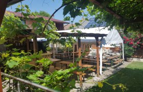 A cozy outdoor area with wooden tables and benches under a leafy canopy, surrounded by plants and flowers.