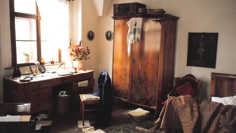 A historic room with a wooden table, sheet music, a wardrobe and a bed. The room looks untidy and old-fashioned.
