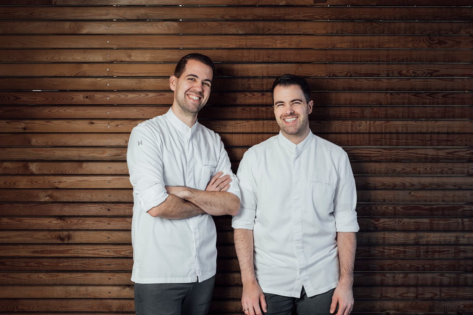 Two men in white shirts stand in front of a wooden wall and smile.