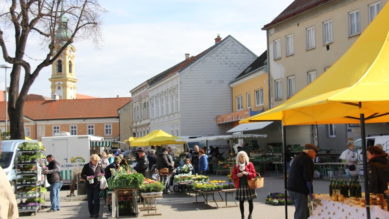 People at a weekly market in Stockerau with yellow market stalls and a church in the background.