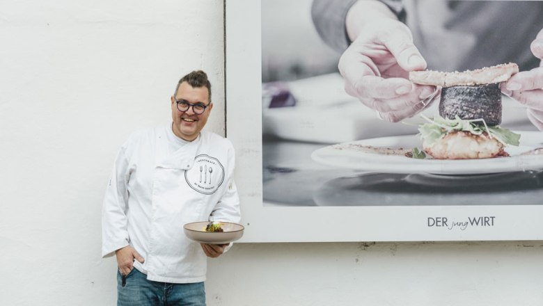 Cook in white jacket with plate in front of poster "DER jungWIRT".
