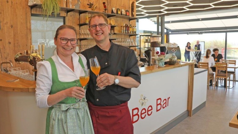 Two people in traditional dress stand smiling in front of the Bee Bar with drinks in their hands.