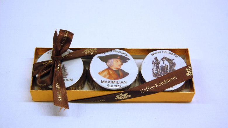 A golden box with three chocolates, decorated with a picture of Maximilian I and Wiener Neustadt motifs, tied with a brown ribbon.