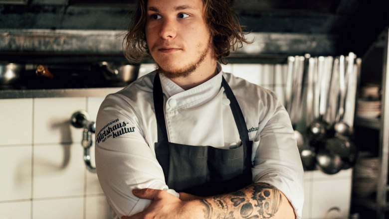 Young chef with tattoos in a professional kitchen, arms crossed.