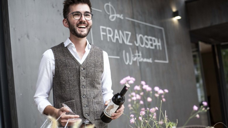 Man with glasses and vest holding wine bottle in front of restaurant 'Franz Joseph'.