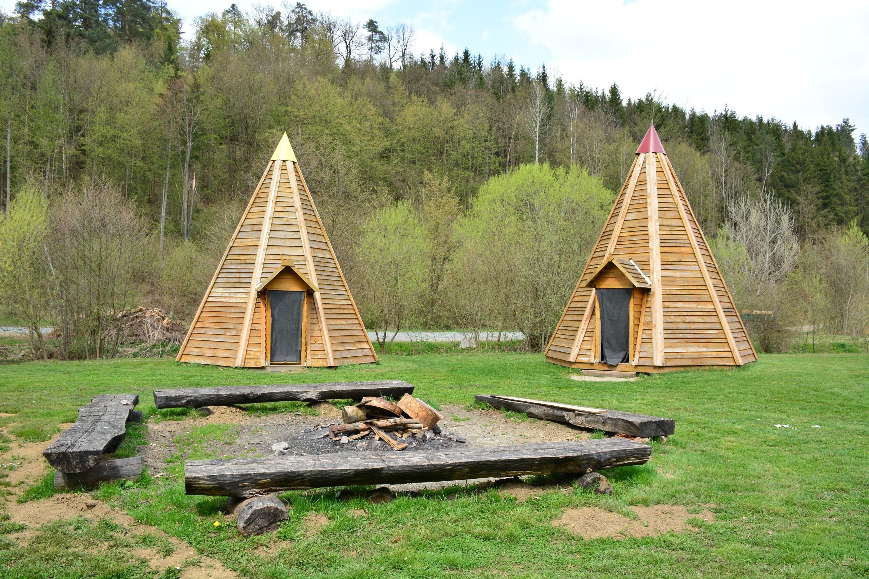 Two wooden tepees on a meadow in front of a forest.