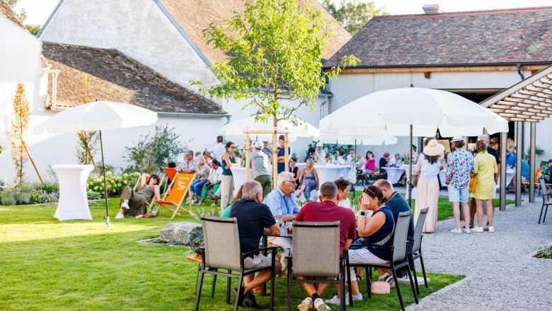 People enjoying an outdoor event with tables, chairs and parasols on a lawn in front of a building.