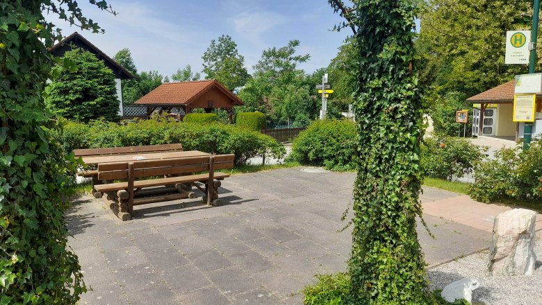 A quiet square with wooden benches and tables, surrounded by green plants and an ivy-covered archway.