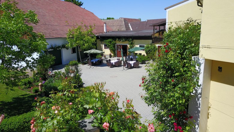 Courtyard of a vineyard with tables, chairs and parasols, surrounded by flowering plants and buildings with red roofs.