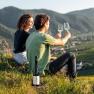 Two people sit on a hill and clink glasses of wine, with a mountain landscape in the background.