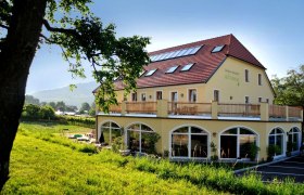 Yellow building with red roof and terrace, surrounded by green landscape.