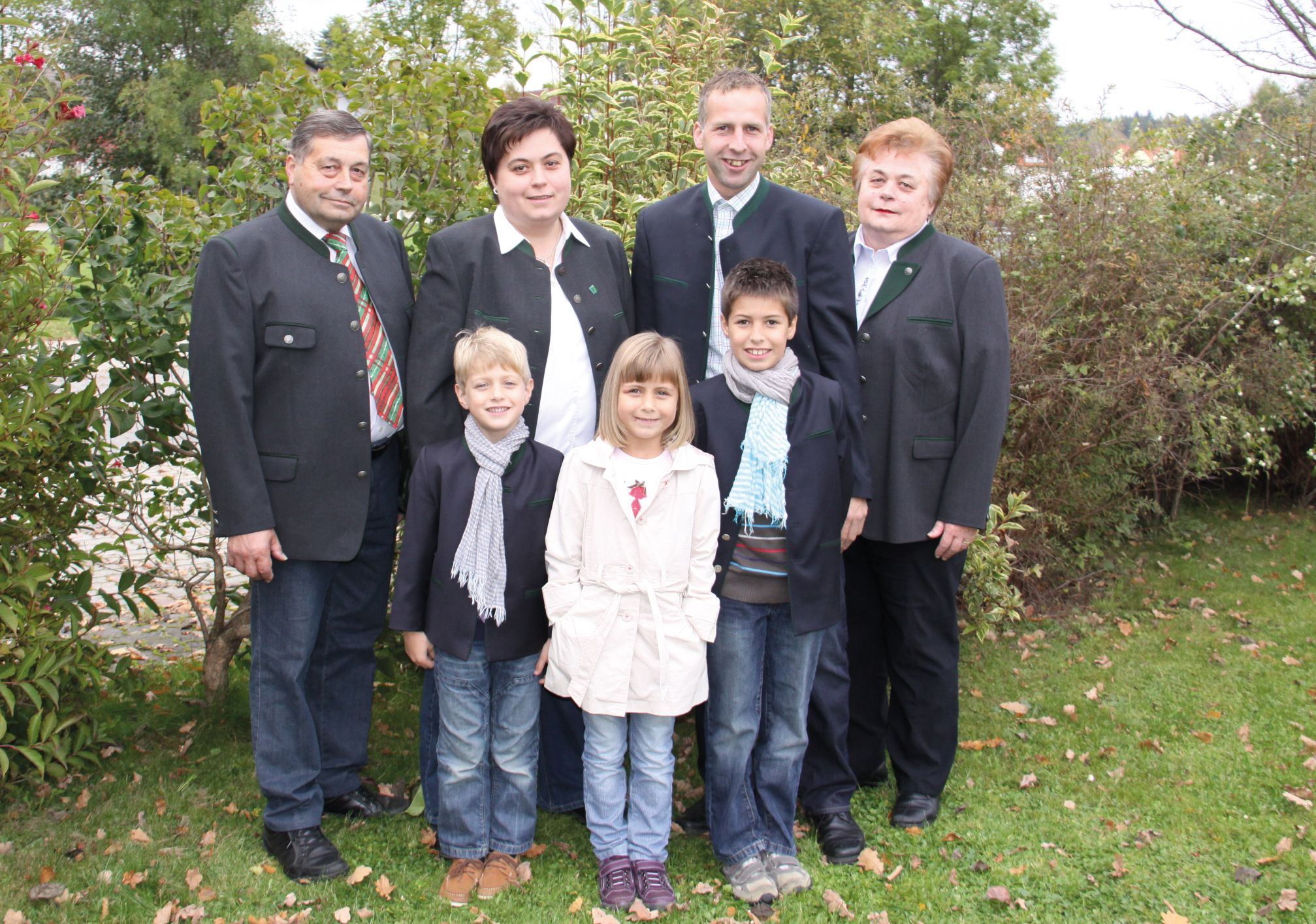 A family is standing outside in a garden, all wearing formal clothes. Bushes and trees can be seen in the background.
