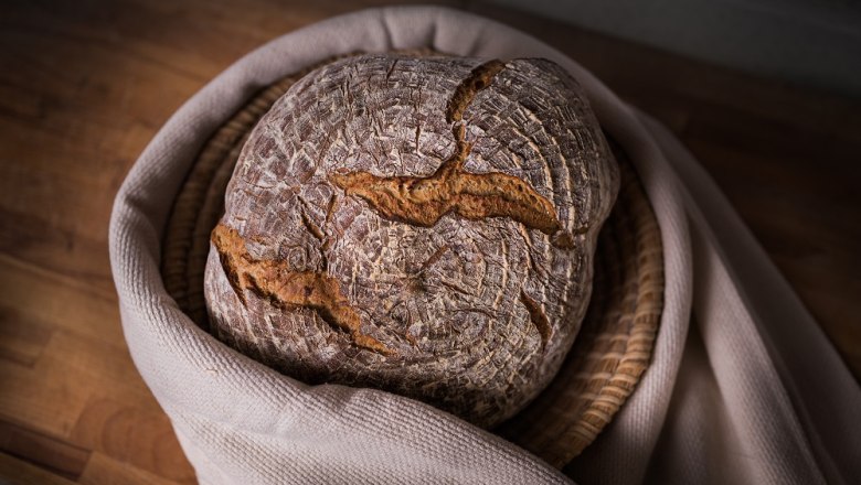 A loaf of bread in a basket on a wooden table, surrounded by a cloth.