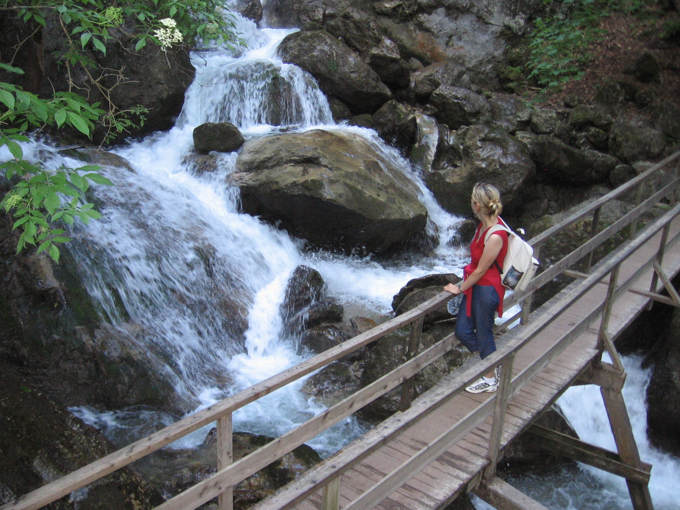 Person on wooden bridge near waterfall in wooded surroundings.