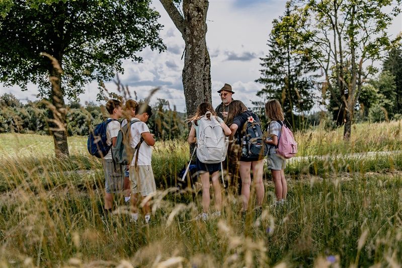 Group of children with backpacks and an adult outdoors, surrounded by trees and grass.