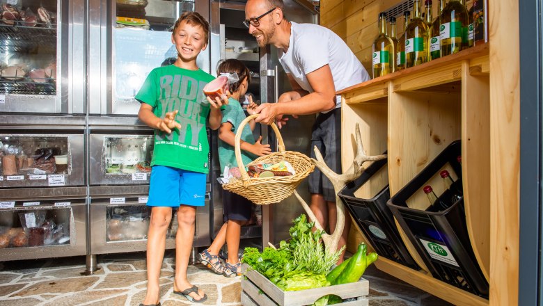 Two children and a man in a store with groceries, including vegetables and bottles.